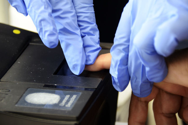 es ist ein Fingerabdruckscanner mit einem Finger, der von zwei Händen geführt wird, abgebildet (verweist auf: Eurodac)