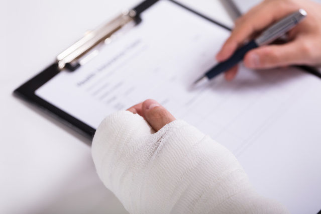 die Abbildung zeigt eine linke Hand in einem Verband und mit der rechten Hand wird ein Formular ausgefüllt (Quelle: Andrey Popov - stock.adobe.com) die Abbildung zeigt eine linke Hand in einem Verband und mit der rechten Hand wird ein Formular ausgefüllt