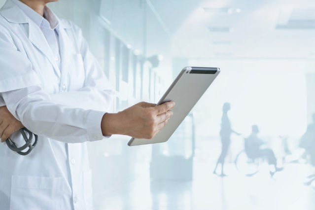 es ist im Vordergrund eine Ärztin mit einem Tablet in der Hand und im Hintergrund eine Person die eine andere Person im Rollstuhl schiebt abgebildet (verweist auf: Krankenhausentlassberichte)