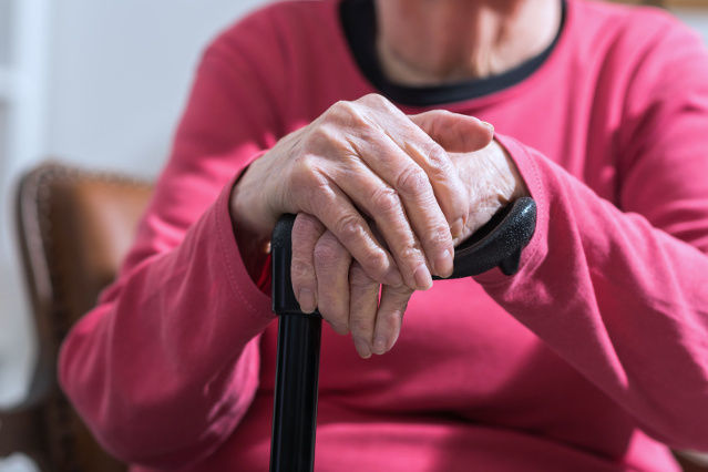 es ist eine ältere Person ab dem Schulterbereich, sitzend mit den Händen auf einem Gehstock, abgebildet