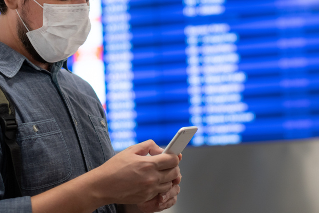 es ist ein Mann mit Mund-Nasenschutz sowie Handy in der Hand abgebildet und im Hintergrund ist eine Flughafenanzeigetafel  (verweist auf: Corona-SMS oder auch Einreise-SMS)