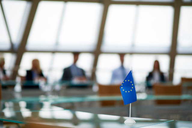 people are sitting at a conference table and a small EU flag is in the foreground on the table